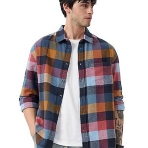 A man with dark wavy hair wearing an open multi-colored checkered flannel shirt in shades of orange, blue, red, and yellow over a plain white t-shirt, paired with light-wash denim jeans.